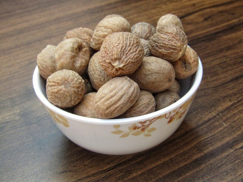 on a dark brown wooden surface is a white bowl with flower prints full of whole nutmegs