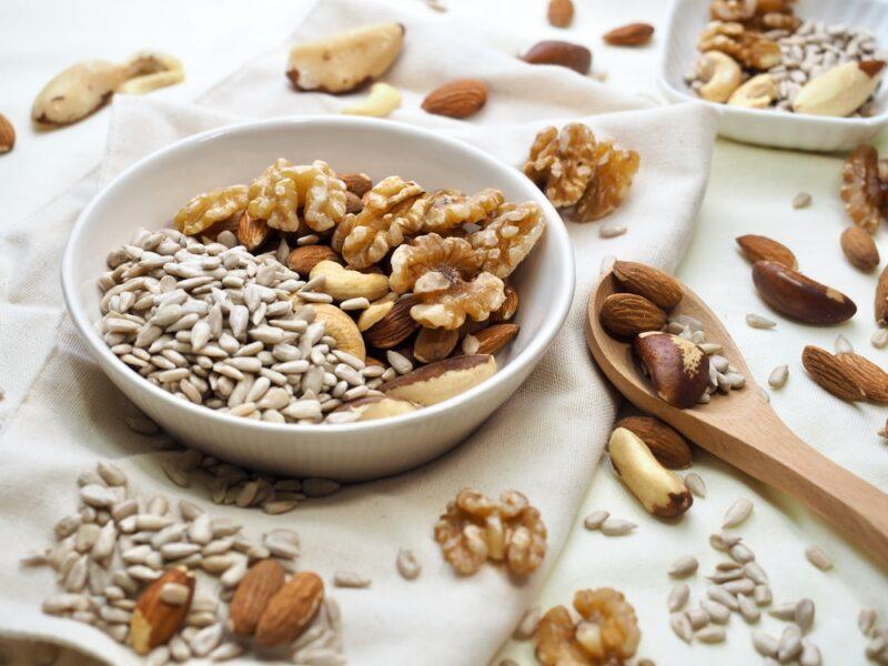 a shallow bowl of nuts and seeds resting on a white table napkin with loose nuts and seeds around it, a wooden spoon lays beside it