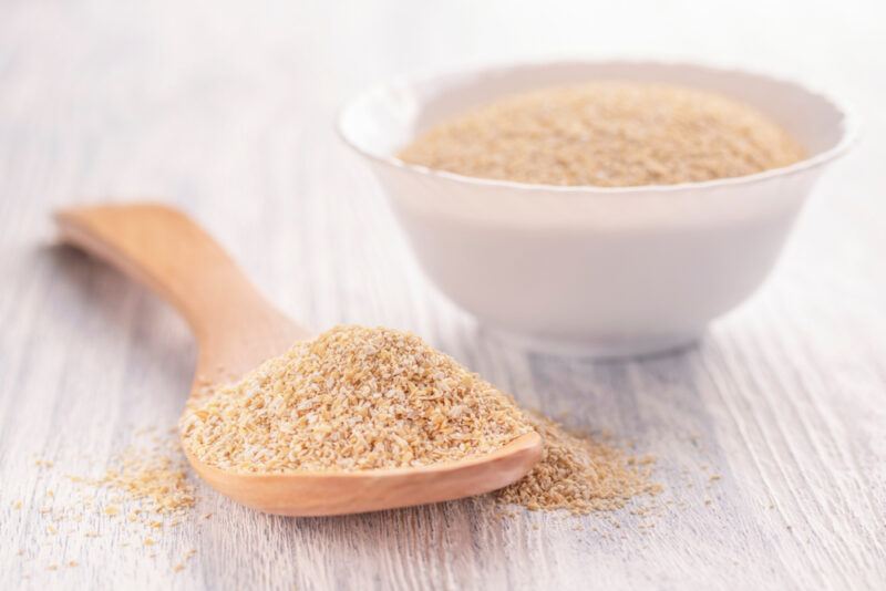 on a light colored wooden surface is a wooden spoon full of oat bran with a white bowl at the back also full of oat bran