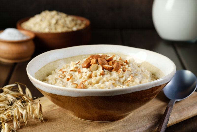 on a wooden chopping board is a ceramic brown and white bowl full of oatmeal with chopped almonds on top and silver spoon beside it, at the back is a wooden bowls of raw oats and sugar, and white ceramic jar