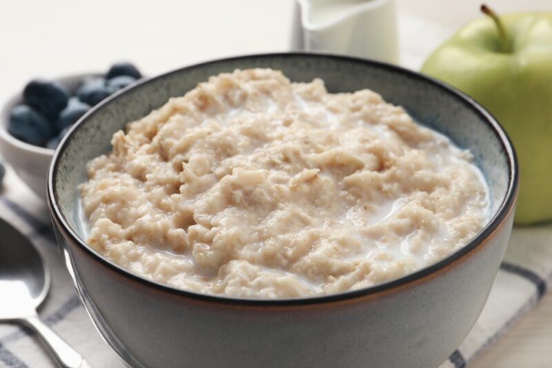  a closeup image of a ceramic bluish bowl with dark blue and gold rim full of oatmeal porridge, at the back is a green apple and a small bowl of blueberries