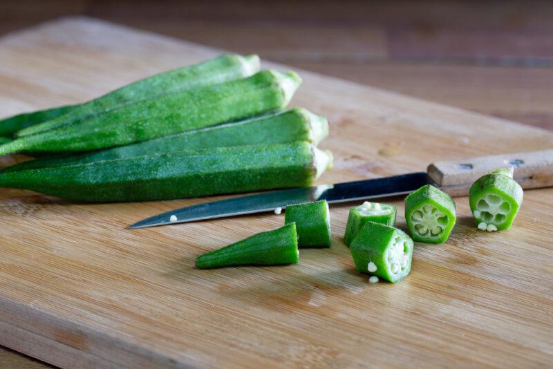 a closeup image of a wooden chopping board with whole okras and chopped okra with a knife