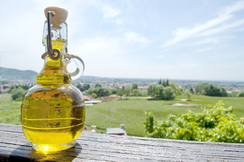 A glass container of light olive oil rests on a wooden table in front of a beautiful green landscape.