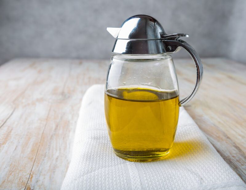 a clear jug with metal handle of olive oil resting on a table napkin on a wooden table