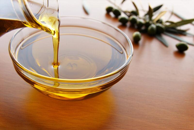 partial image of a glass jar of olive oil pouring its contents onto a clear glass bowl, on a brown wooden surface with a few loose olives and bunch of olives behind it