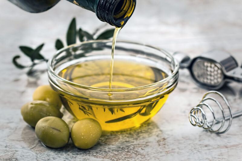 on a marble surface is a clear bowl of olive oil with whole olives around it, beside is a metal mixer and metal strainer