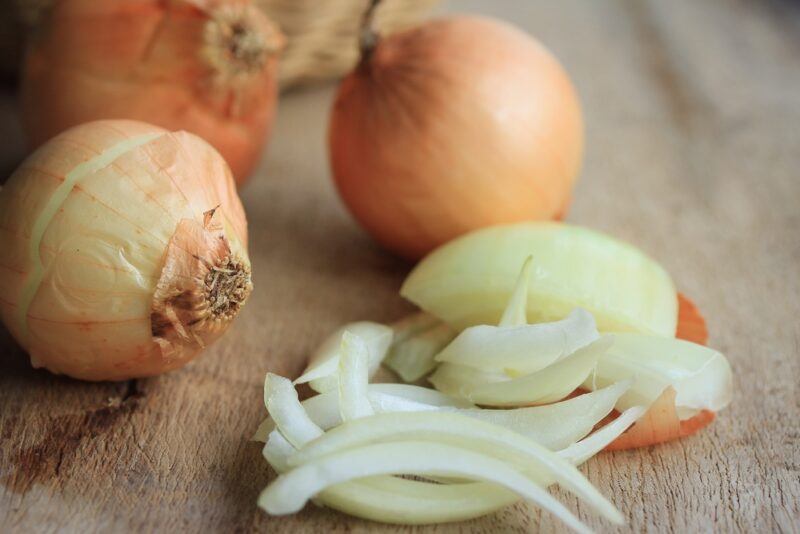 a closeup image of three whole onions and a sliced one on a wooden chopping board