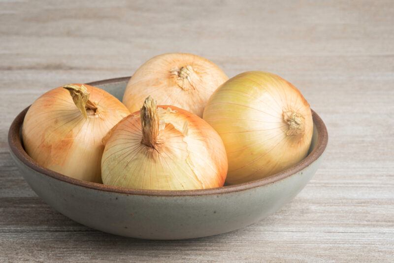 on a wooden surface is a tan colored ceramic bowl with four white onions