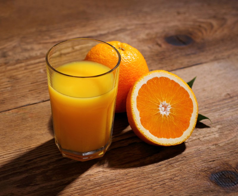 A whole orange and a half orange rest next to a glass of orange juice on a rough wooden background.