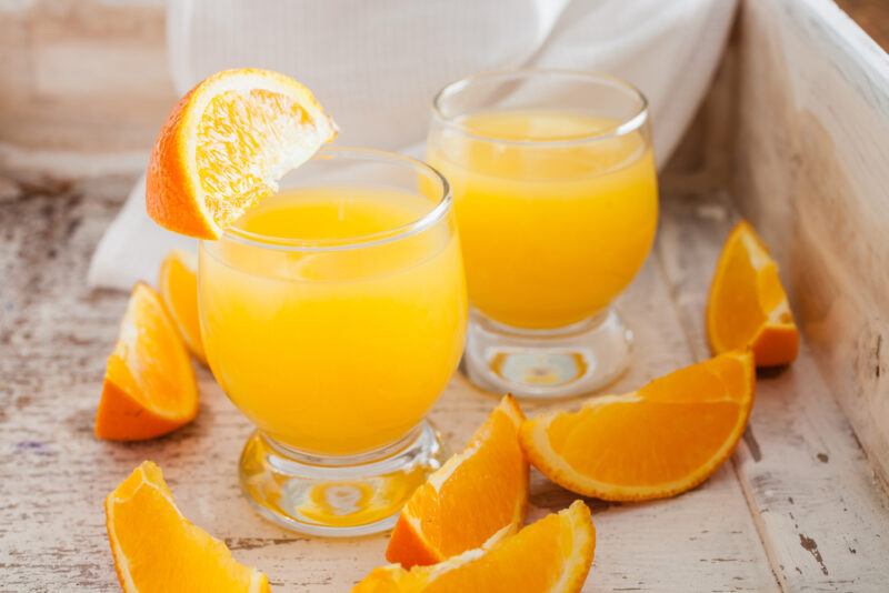 on a wooden tray is a couple of clear glasses with orange juice, with orange loose wedges around the glasses