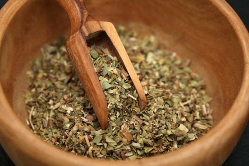 a closeup image of a wooden bowl with dried oregano leaves and a wooden scoop shove in it