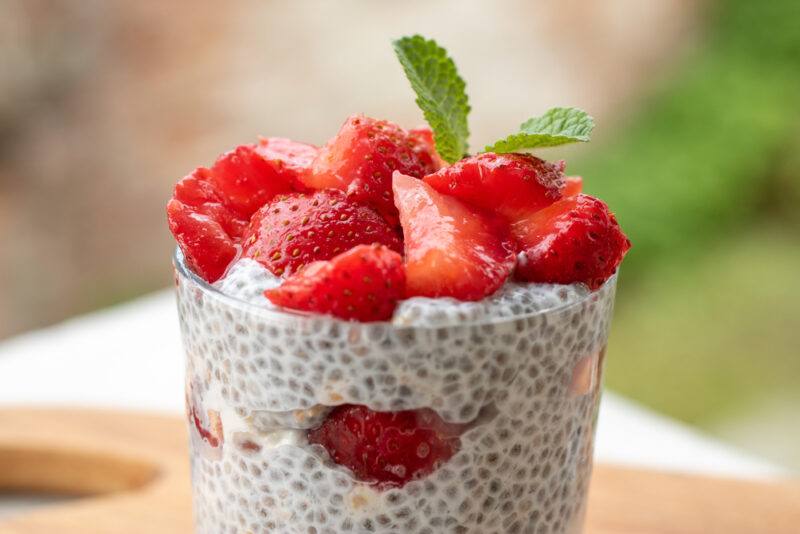 a closeup image of a clear glass of overnight coconut chia pudding with sliced strawberries on top with a sprig of herb