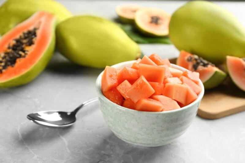 on a concrete surface is a white bowl of cubed papaya and silver spoon, with halve and whole papayas at the back