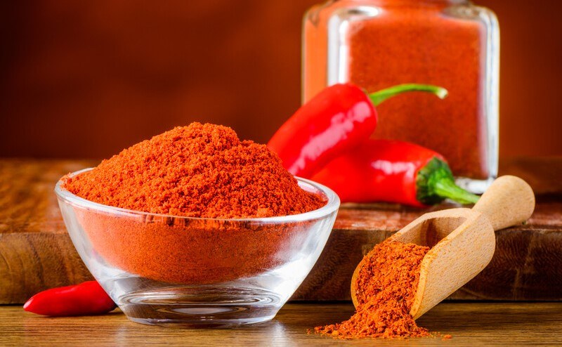 a white glass bowl with paprika powder, beside it is a wooden scoop with paprika powder, at the back is a couple of fresh paprika with a glass jar full of paprika powder 