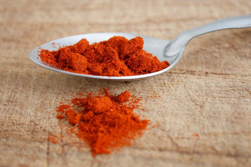 closeup image of a metal spoon full of paprika with loose paprika powder on the wooden surface