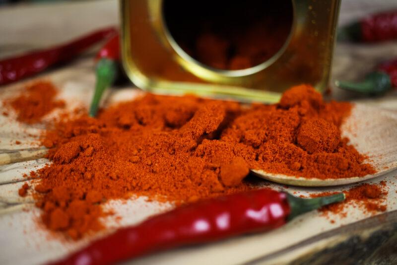 closeup image of a mound of paprika poured from a topped tin container, a fresh chili pepper can be seen at the foreground and a few more at the background