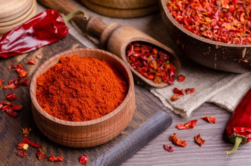 a wooden bowl full of paprika powder resting on top of an aged wooden chopping board, with loose dried chili flakes beside it, at the back is a wooden scoop and wooden bowl at the back with dried chili as well