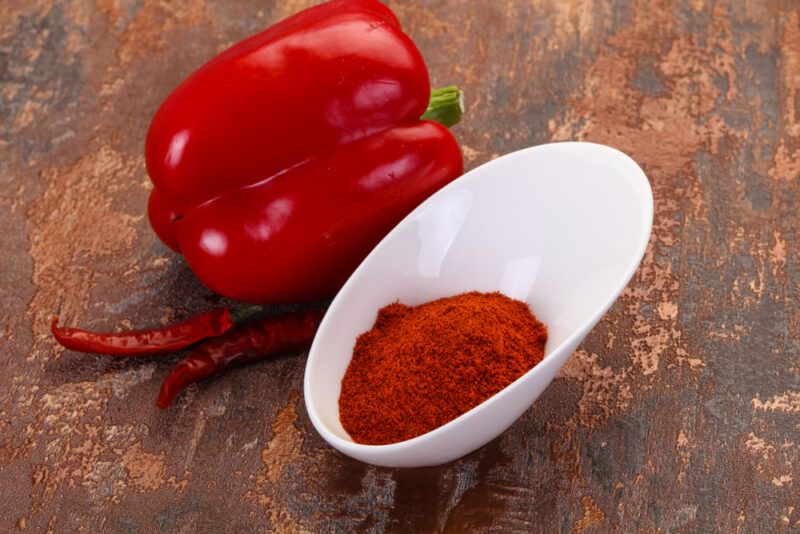 on a rustic looking surface is a white odd-shaped bowl with paprika powder, behind it is a whole bell pepper and a couple of red chili peppers