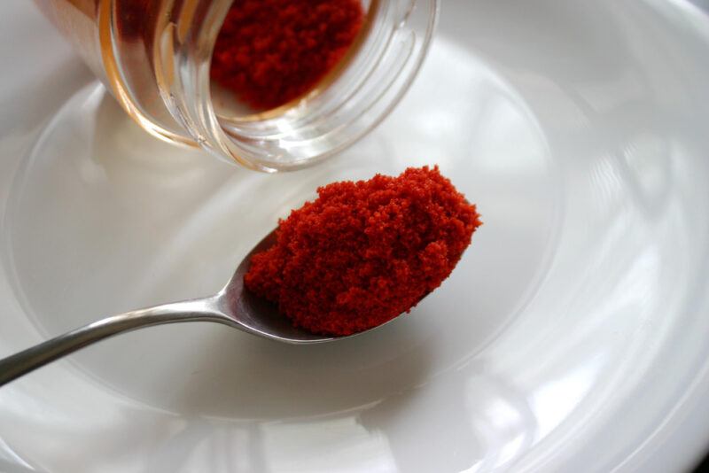 a closeup image of a white plate with a silver spoon full of paprika, beside it is the jar of paprika powder