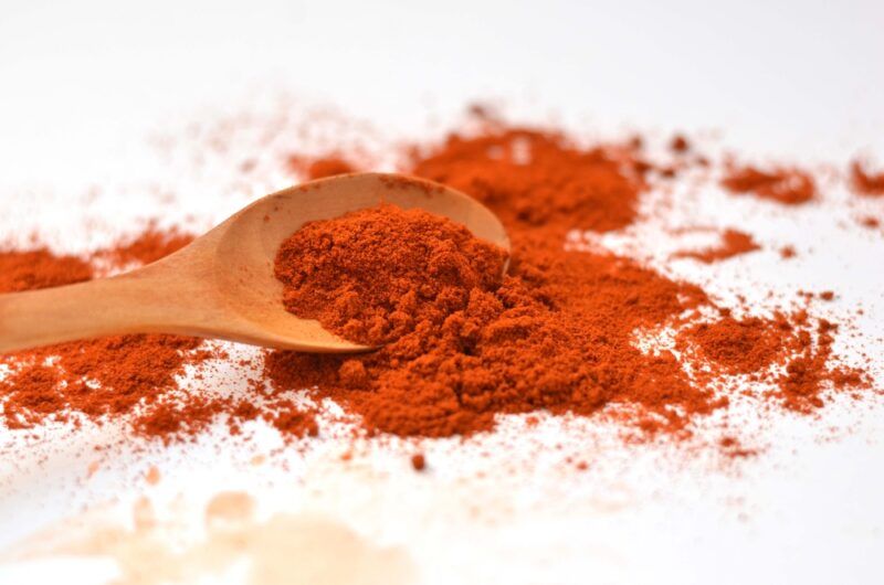 on a white surface is a wooden spoon with paprika powder, around it are loose paprika powder as well