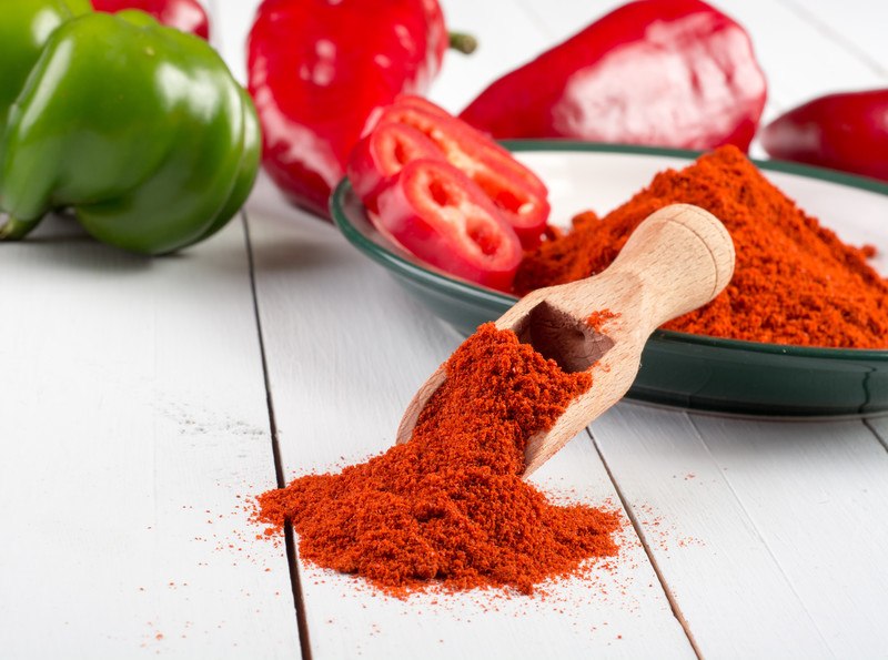  a wooden scoop full of paprika powder with a mound spilled over the white wooden surface, its handle is resting on a plate with paprika powder and sliced paprika, at the back are fresh paprika peppers