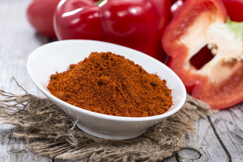 a white cylindrical odd shaped-bowl full of paprika resting on a burlap sack with fresh peppers at the back