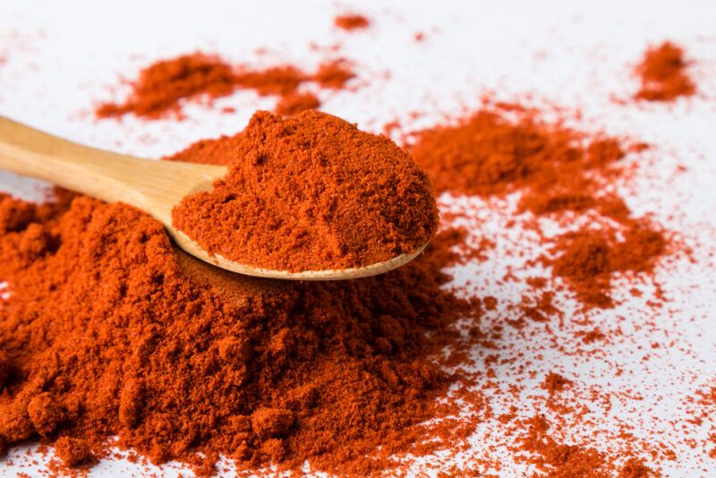 on a white surface is a closeup image of a mound of paprika powder with a wooden spoon on top also with paprika powder