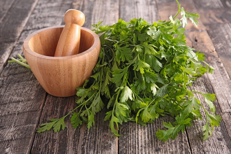 on an aged wooden surface is a bunch of fresh parsley with a wooden mortar and pestle beside it