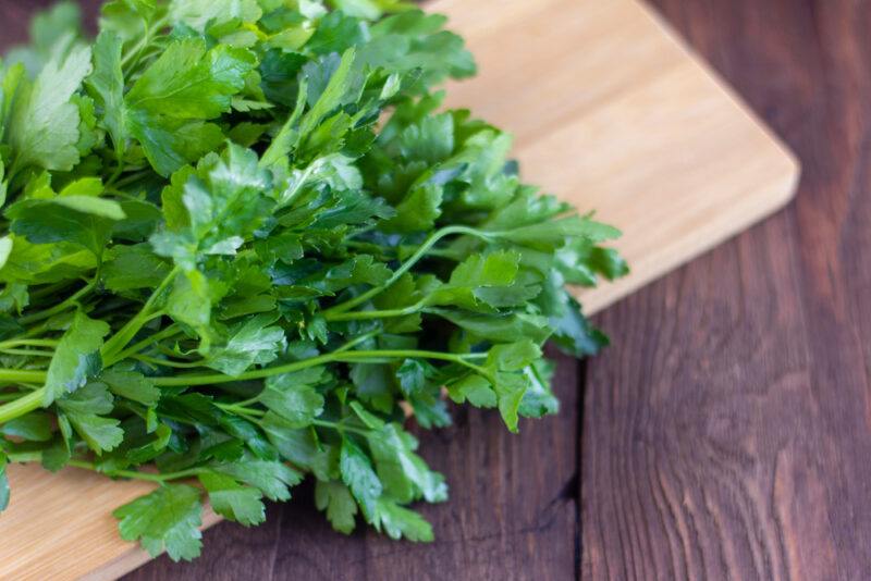 on a rustic looking wooden surface is a light brown wooden chopping board with a bunch of fresh parsley