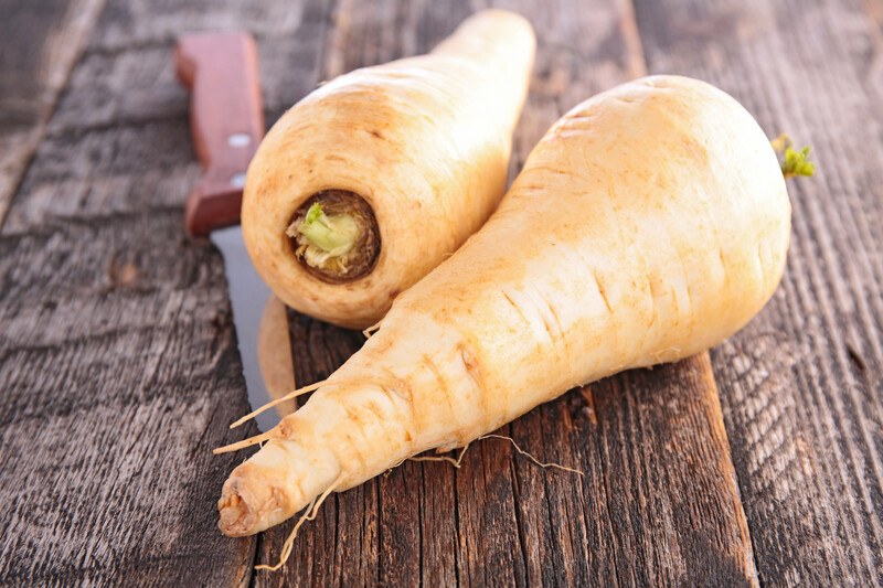 on a rustic wooden surface is a couple of whole parsnips with a knife beside it