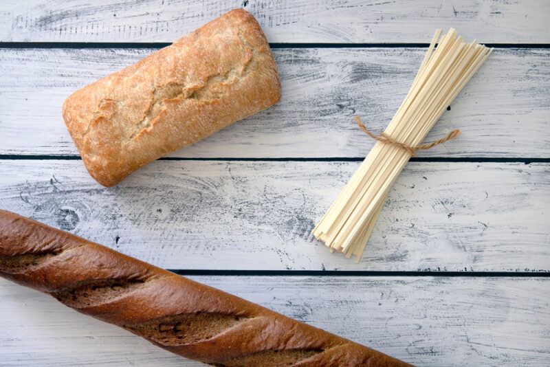 on a faded white wooden surface is an overhead image of fresh bread and pasta 