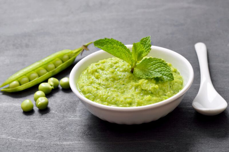 on a dark grey concrete surface is a white bowl with pea puree with herbs on top, beside it are fresh peas with pods beside it and a white ceramic spoon