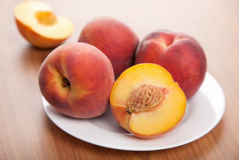 on a wooden surface is a white plate with four peaches, one cut open; at the back is a halved peach