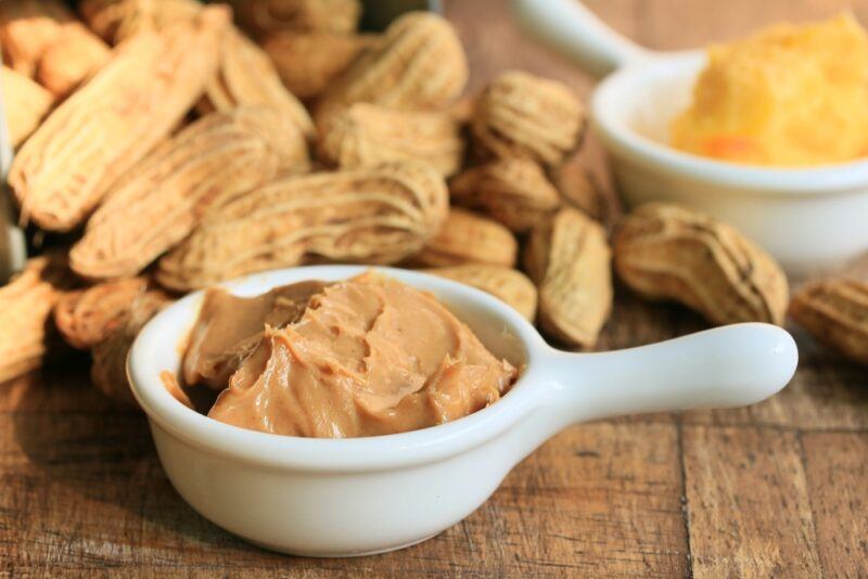 on a wooden surface is a white ceramic bowl with handle full of peanut butter, at the back is a pile of fresh peanuts