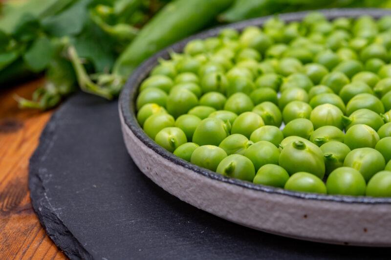 on a wooden surface is a closeup image of a dish with peas resting on a black stone board with pea pods at the back