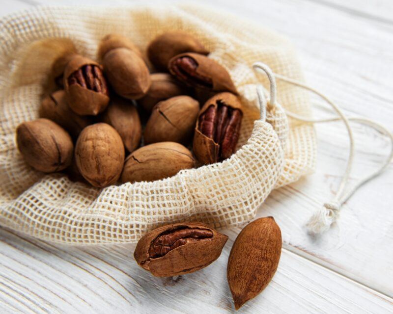 on a white wooden surface is a partially open cloth string bag with pecans, a couple of pecan nuts beside it