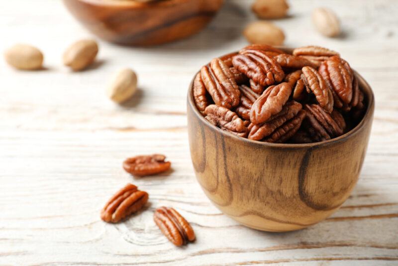 on a white wooden surface is a small wooden bowl full of pecans, with loose pecans around it