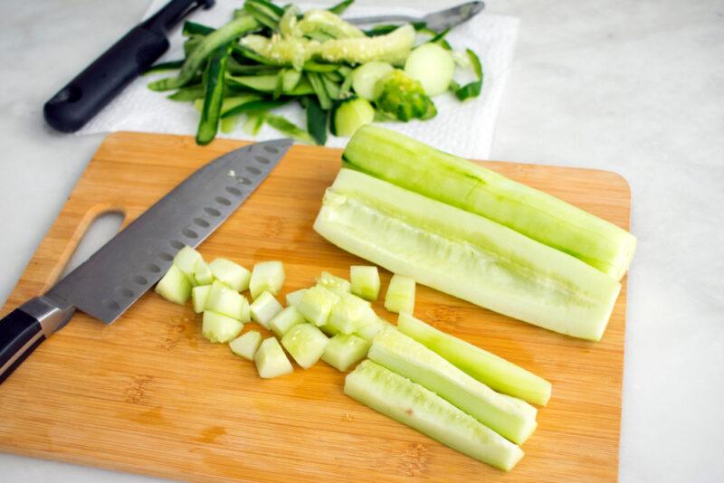 on a marbled surface is a wooden chopping board with peeled and sliced cucumber and a knife on it, at the back are peeled skins and seeds of the cucumber with a peeler beside it