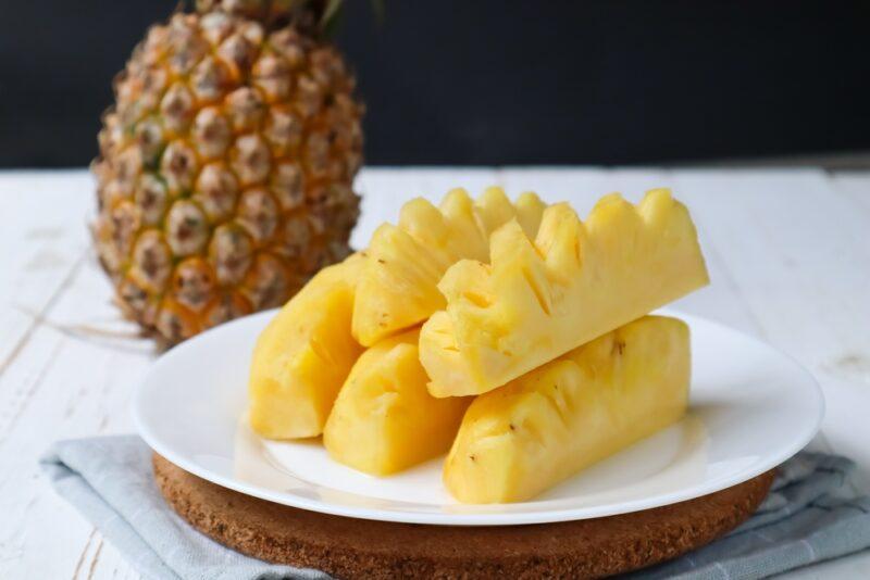 on a marble surface is a white plate with pineapple slices resting on a wooden chopping board, at the back is a whole pineapple