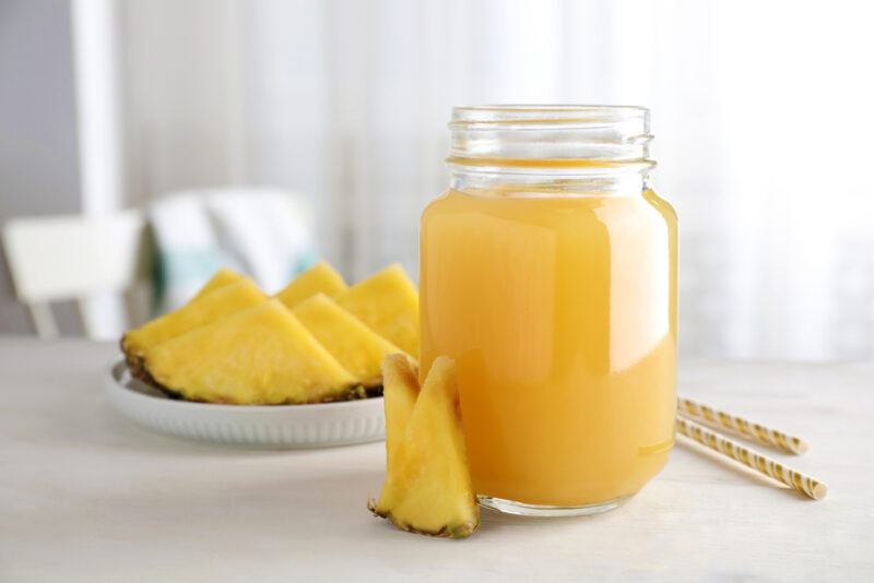 a mason jar of pineapple smoothies with a sliced of fresh pineapple beside it and yellow and white straws, at the back is a white plate with sliced fresh pineapples