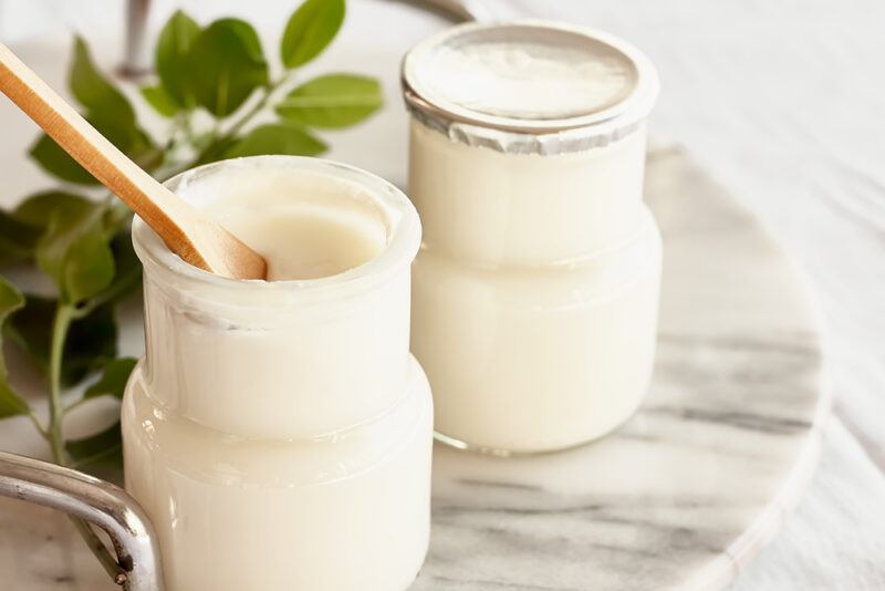 on a round marble board is a couple of jars with plain yogurt, one jar covered with aluminum foil, the other already oven with a wooden spoon in it, at the back is a bunch of green leaves