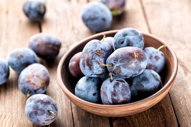 on a wooden surface is a wooden bowl full of plums with loose plums around it