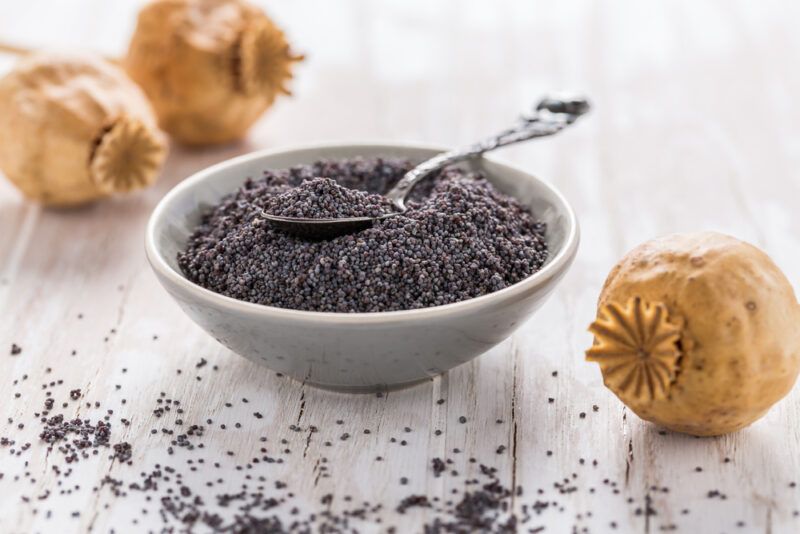 on a white wooden surface is a grey ceramic bowl full of poppy seeds with loose poppy seeds and poppy heads around it