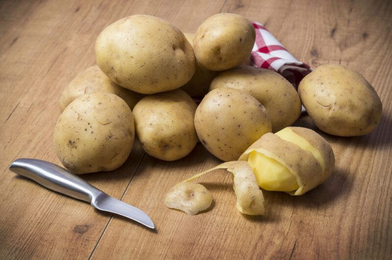 on a wooden surface is a pile of potatoes with one that is partially peeled with a knife beside it