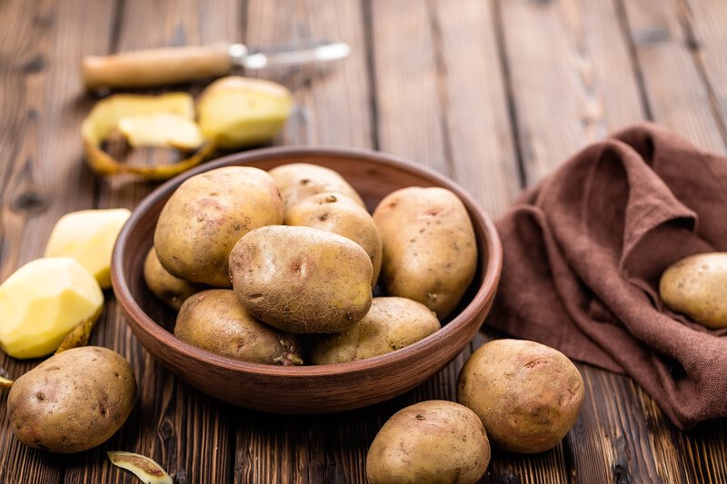 on an aged wooden surface is a a wooden bowl full of potatoes, with loose potatoes around it and a vegetable peeler and brown table napkin at the back