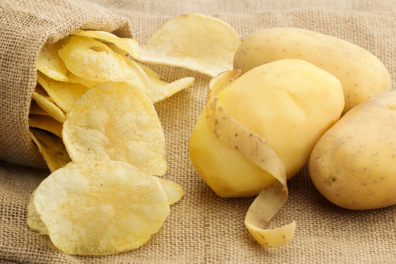 on a burlap sack is a closeup image of pealed whole potato and a couple of whole potatoes with skin still on,  and potato chips pouring out of a burlap pouch