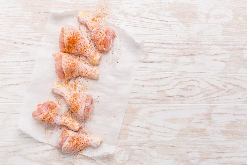 on a wooden surface are chicken drumsticks with poultry seasoning laid out on a white parchment paper