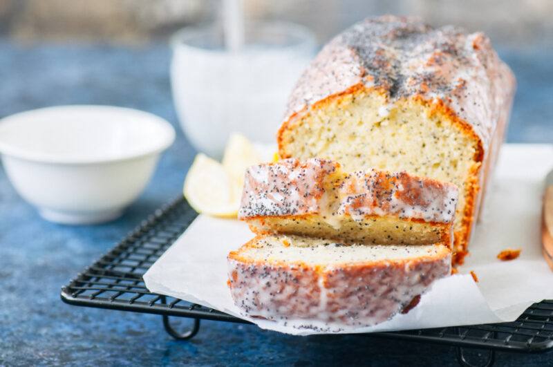 partially sliced pound cake resting on a cooling rack with white baking paper underneath, at the back is a small ceramic bowl and cup 