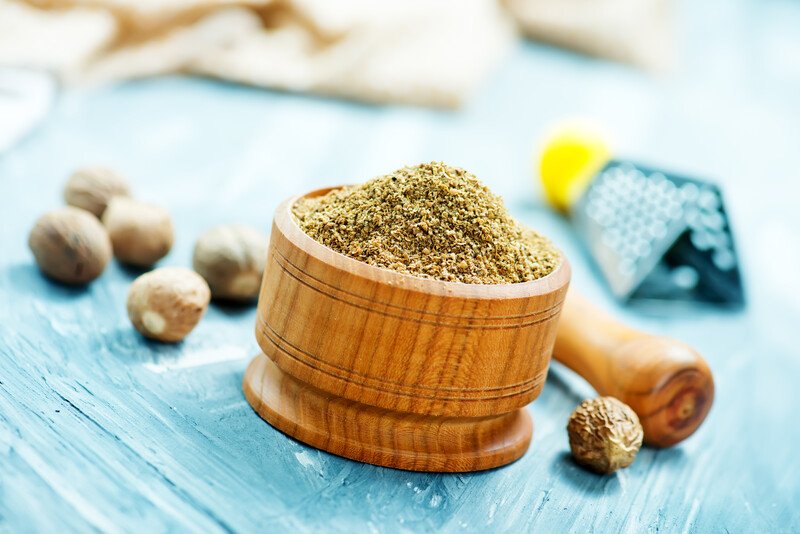 on a blue wooden surface is a wooden mortar and pestle full of ground nutmeg with whole nutmegs and metal grater at the back