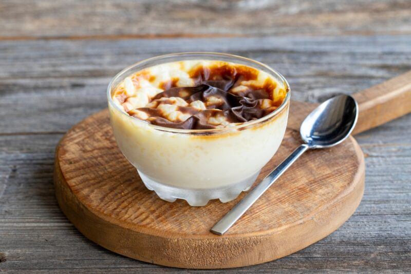 on a rustic wooden surface is a round wooden board with handle with a glass bowl of milk pudding on it and a silver spoon beside it